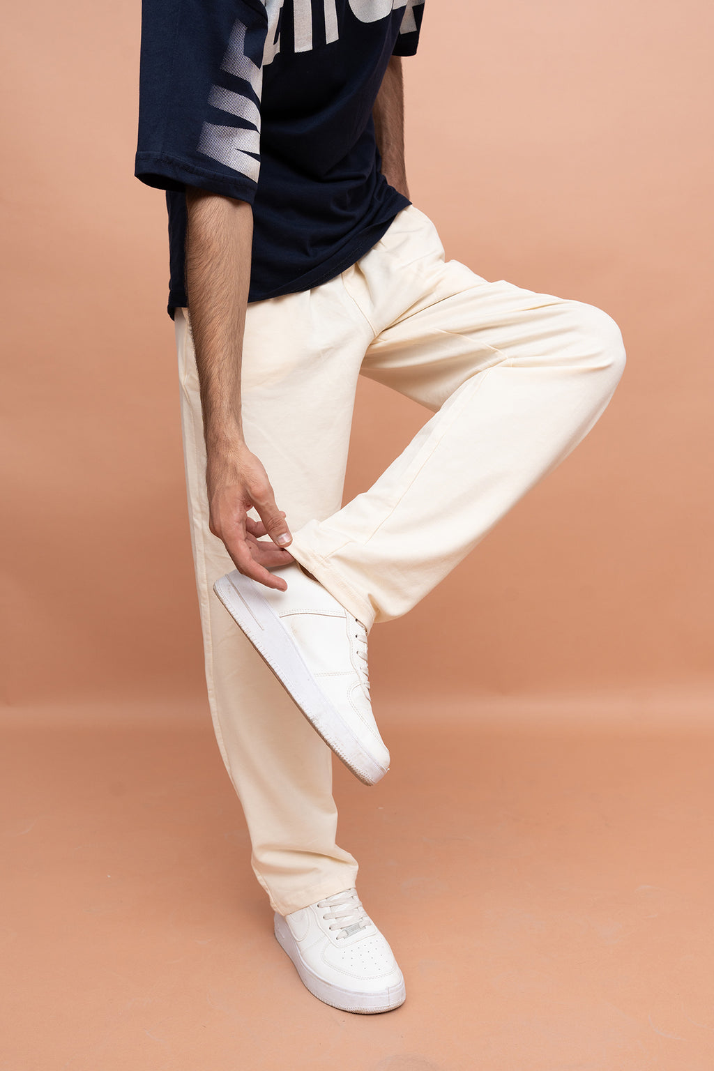 A person wearing cream-colored relaxed-fit pants, a navy graphic t-shirt, and white sneakers, standing against a solid peach-colored studio background.