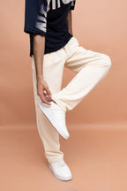 A person wearing cream-colored relaxed-fit pants, a navy graphic t-shirt, and white sneakers, standing against a solid peach-colored studio background.